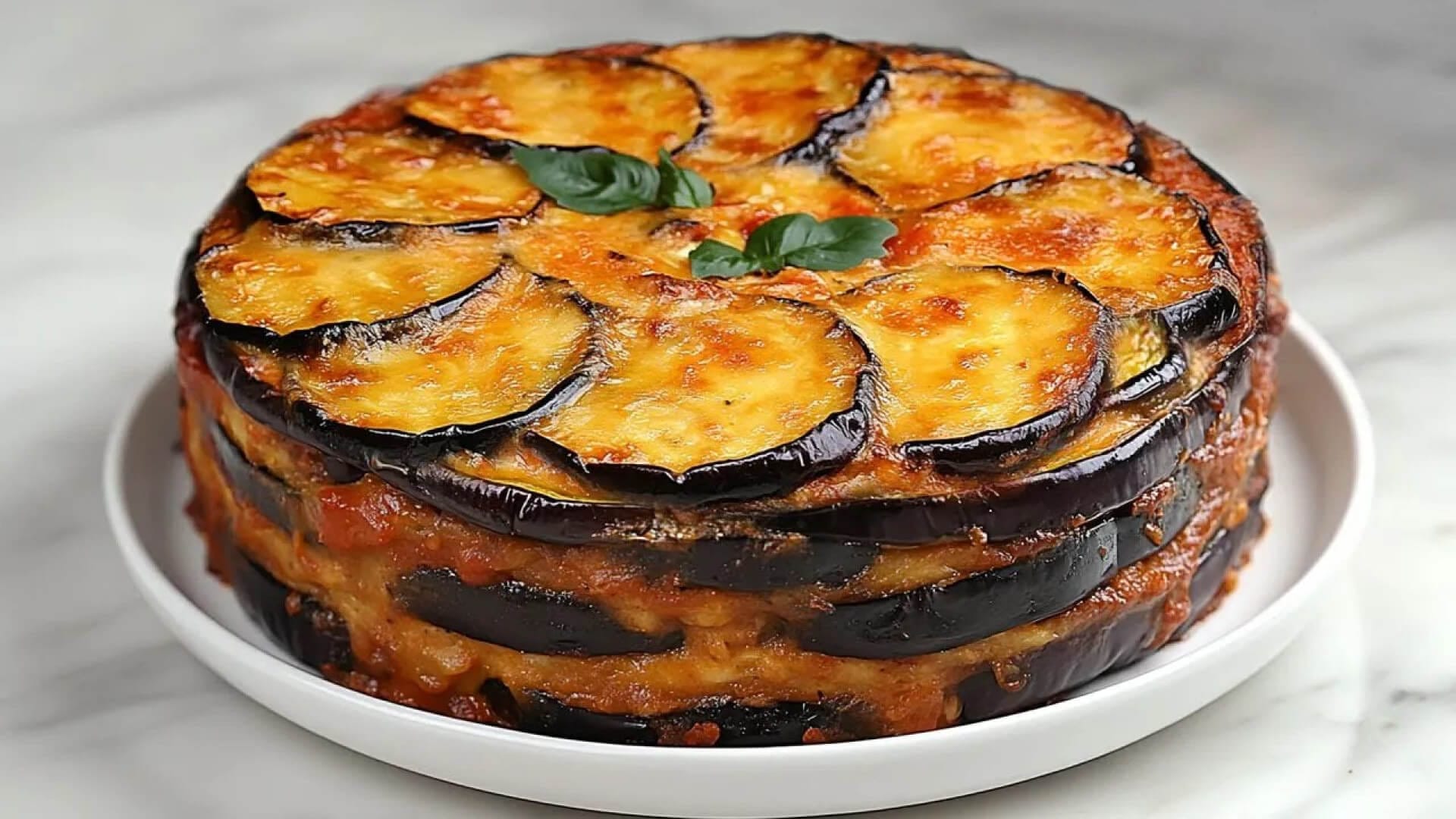 طرز تهیه گراتن بادمجان بدون گوشت به صورت قالبی و چند لایه که روی آن با پنیر طلایی و برگ ریحان تزیین شده است.