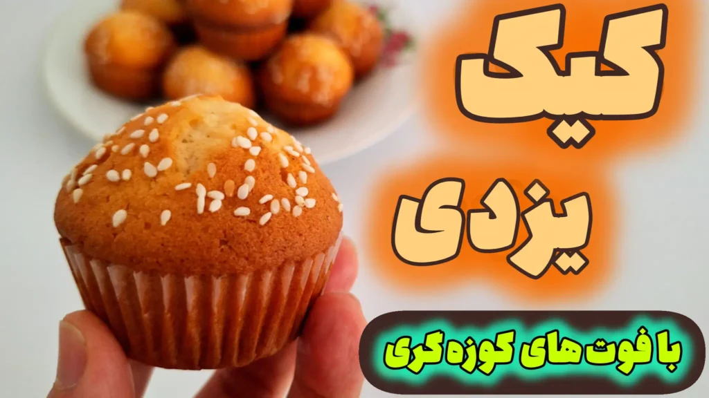 دستی که یک عدد کیک یزدی خانگی طلایی و گنبدی شکل با رویهی کنجدی را نگه داشته است. در پسزمینه، بشقابی پر از کیکهای یزدی تازه پخته شده دیده میشود.