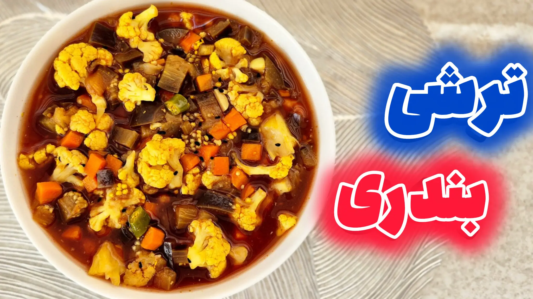 نمای نزدیک از یک کاسه سفید پر از ترشی بندری اصل خانگی، شامل تکههای گل کلم، هویج، بادمجان و فلفل در سس غلیظ و قرمز رنگ.