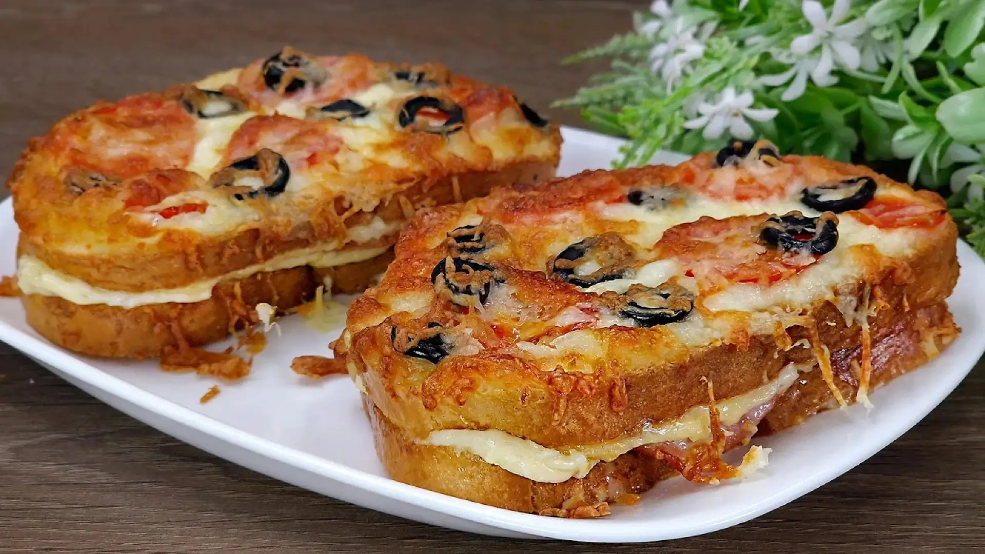 دو عدد ساندویچ پیتزا با نان تست ضخیم در یک بشقاب سفید. روی ساندویچ‌ها با پنیر طلایی، گوجه و زیتون سیاه پوشانده شده و لایه‌ای پنیر از بین نان‌ها پیداست.