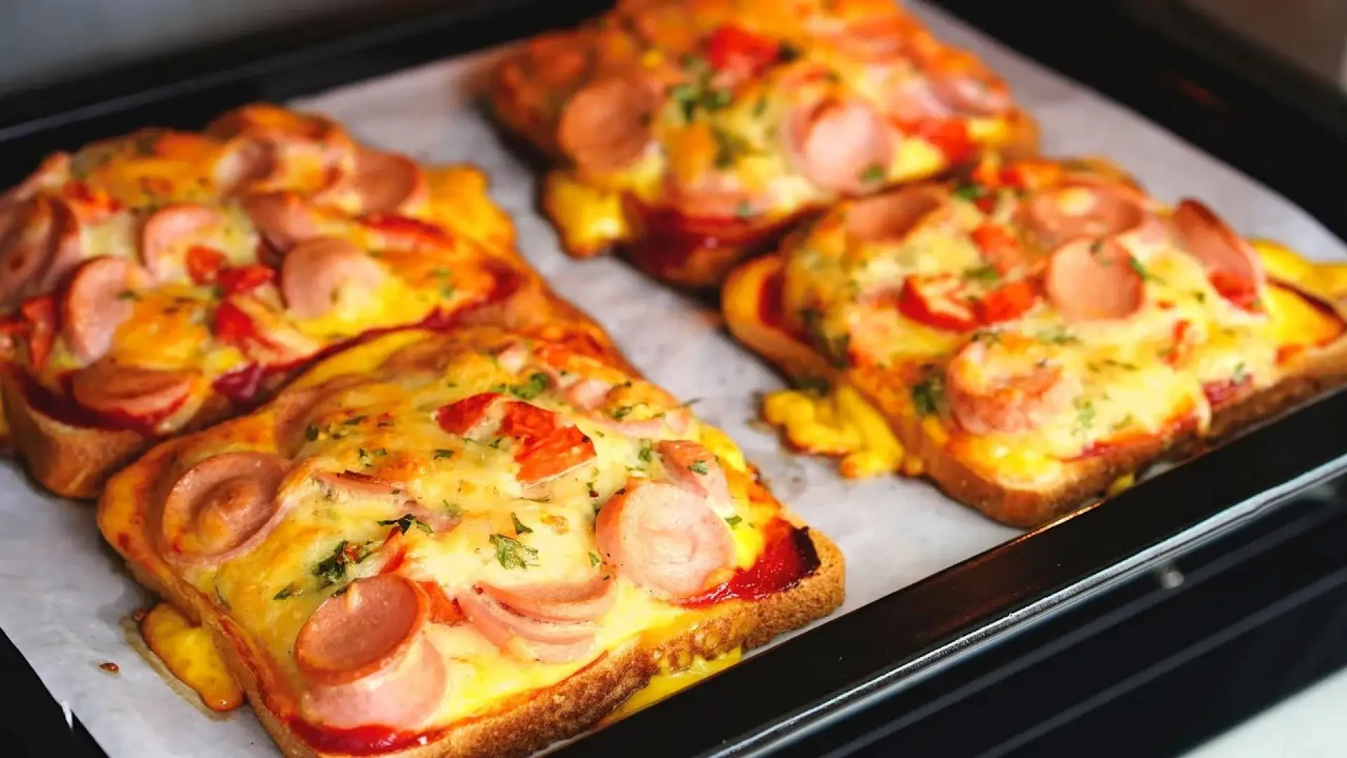 چهار عدد تست پیتزایی با سوسیس حلقه شده، پنیر پیتزای ذوب شده و سبزیجات، روی کاغذ روغنی در سینی فر قرار گرفتهاند.