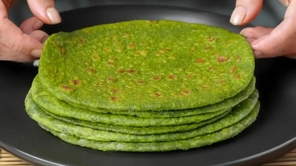 دستهای نان اسفناج گرد و خانگی با رنگ سبز روشن که روی یک بشقاب مشکی چیده شده و دو دست در حال برداشتن نان بالایی هستند.