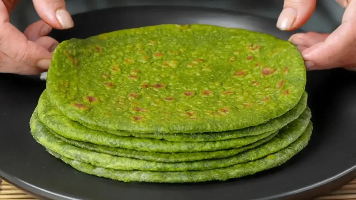 دستهای نان اسفناج گرد و خانگی با رنگ سبز روشن که روی یک بشقاب مشکی چیده شده و دو دست در حال برداشتن نان بالایی هستند.