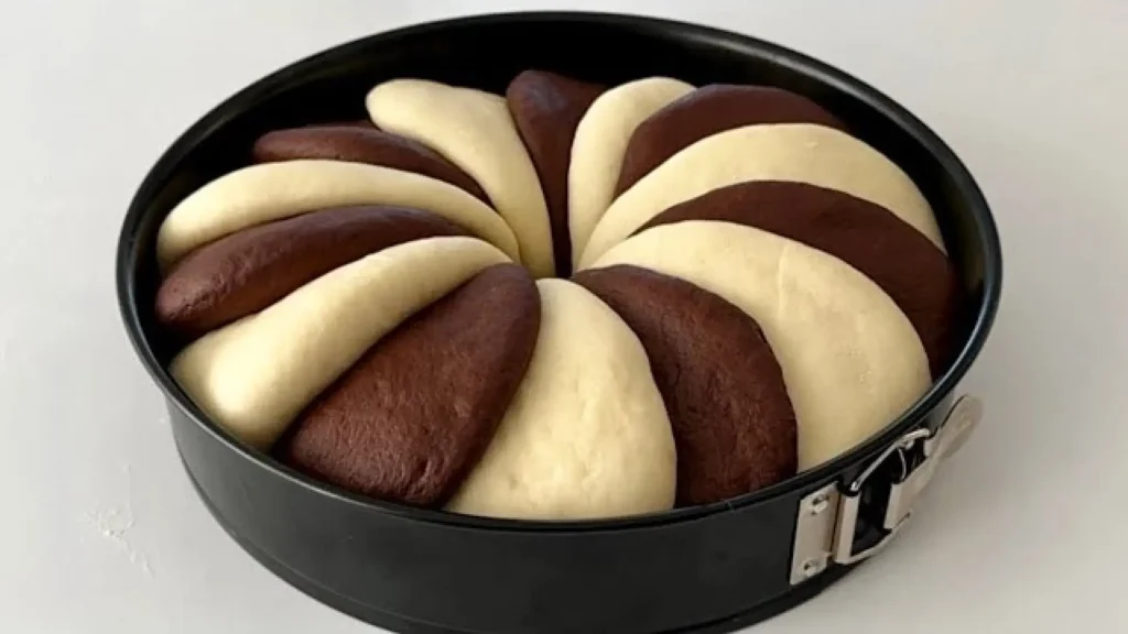 خمیر نان دو رنگ شکلاتی که به صورت قطعات هلالی و یکی در میان در قالب کمربندی مشکی چیده شده و آماده پخت است.