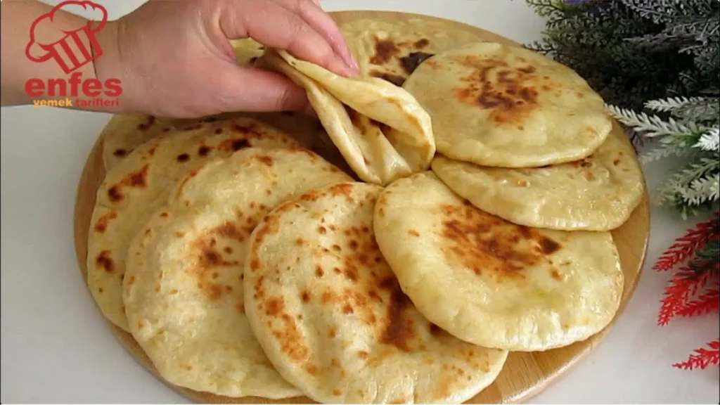 چند نان پیتای گرد و خانگی با لکه‌های طلایی-قهوه‌ای که روی یک تخته چوبی چیده شده‌اند و یک دست در حال تا کردن یکی از نان‌ها برای نشان دادن بافت نرم و لایه داخلی آن است.