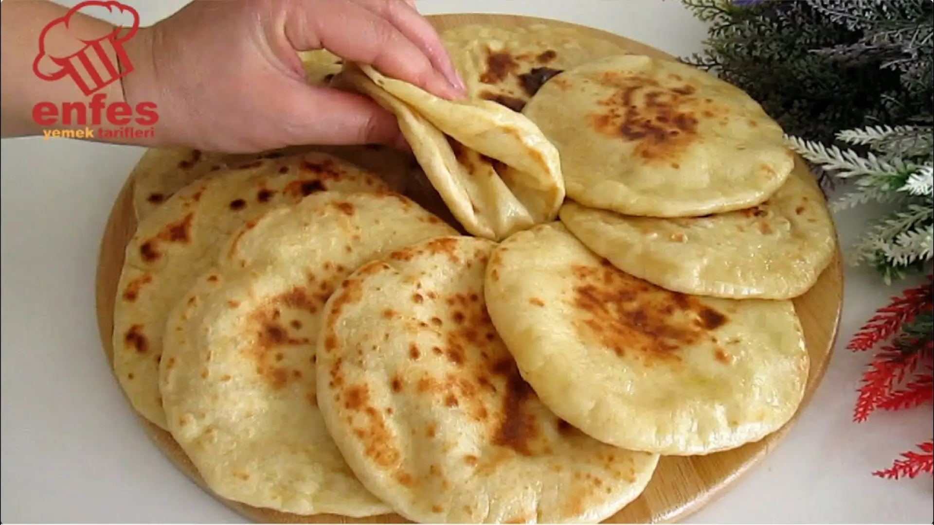 چند نان پیتای گرد و خانگی با لکه‌های طلایی-قهوه‌ای که روی یک تخته چوبی چیده شده‌اند و یک دست در حال تا کردن یکی از نان‌ها برای نشان دادن بافت نرم و لایه داخلی آن است.