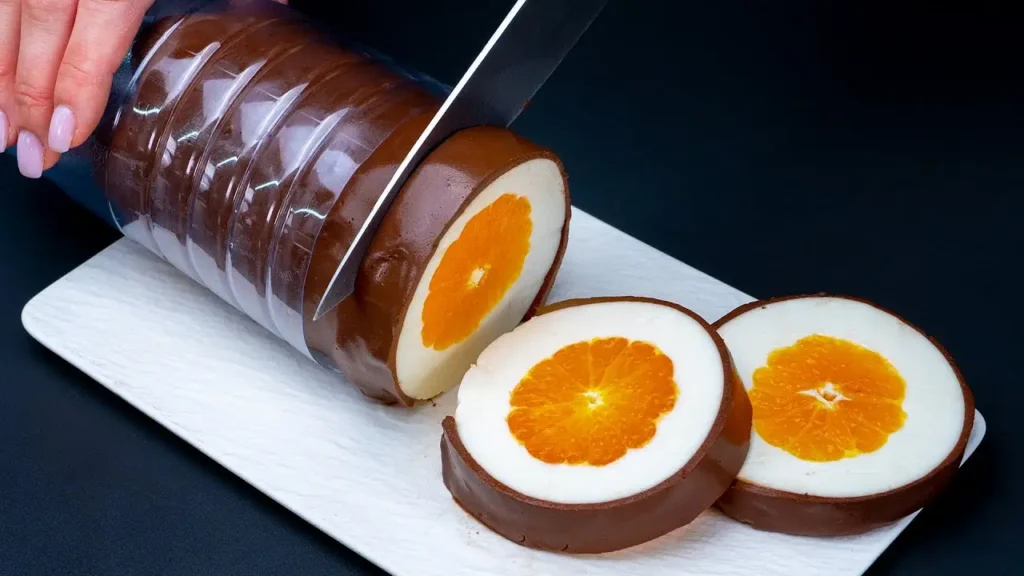 برش زدن دسر نارنگی با شکلات به شکل رولی با مرکز نارنگی تازه و لایه‌های ژله‌ای سفید و کاکائویی